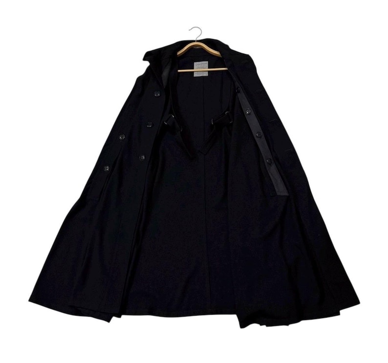 May include: A black trench coat with a wide, open design, revealing the interior lining and button details. The coat is hanging on a wooden hanger, showcasing its full length and tailored silhouette. The coat has a classic, timeless style.