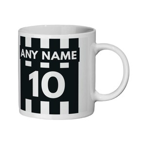 May include: White ceramic mug with a black and white design. The design includes the text "ANY NAME" above the number "10". The mug has a curved handle and a cylindrical shape. The design is reminiscent of a sports jersey.