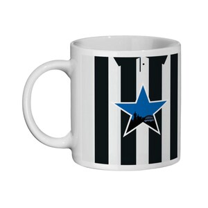 May include: White ceramic mug with black and white vertical stripes. A blue star with a silhouette of a city skyline is centered on the mug. The mug has a curved handle.