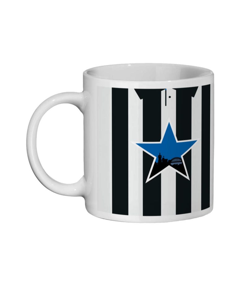 May include: White ceramic mug with black and white vertical stripes. A blue star with a silhouette of a city skyline is centered on the mug. The mug has a curved handle.