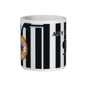 May include: White ceramic mug with black vertical stripes, featuring a team logo and the text "ANY NAME" and the number "9". The mug has a blue accent line and a gold and blue team logo.