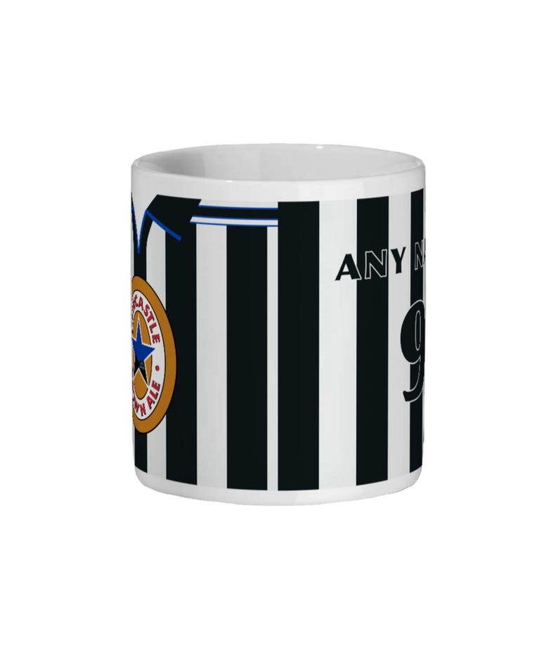 May include: White ceramic mug with black vertical stripes, featuring a team logo and the text "ANY NAME" and the number "9". The mug has a blue accent line and a gold and blue team logo.