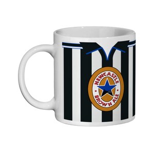May include: White ceramic mug with a black and white striped design, resembling a football shirt. The mug displays a circular emblem with a blue star and the words "Newcastle Brown Ale".