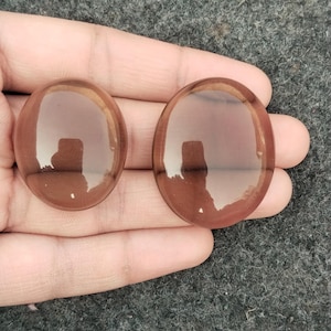 May include: Two oval-shaped, translucent, brown gemstones are displayed. The stones are smooth and polished, with a slight sheen. The background is a textured, dark gray surface. The gemstones are held in a hand.