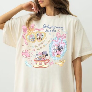 Może przedstawiać: Oversizowy t-shirt w kolorze kremowym z nadrukiem graficznym i napisem "Girls just wanna have fun". Wz&oacute;r zawiera ilustracje Myszki Minnie i Kaczki Daisy, lustra w kształcie serc, filiżankę, szklany pantofelek oraz złotą lampę.