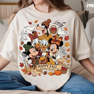 May include: An ivory t-shirt featuring a graphic of Disney characters including Mickey Mouse, Minnie Mouse, Goofy, Donald Duck, Daisy Duck, and Pluto. The design includes autumn leaves, acorns, pumpkins, baskets, and the text "HAPPY FALL Autumn Season".