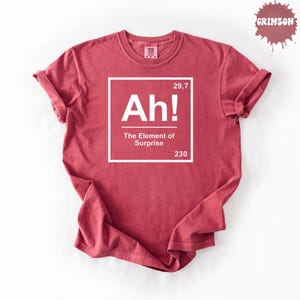 May include: A crimson red short-sleeve t-shirt featuring a white square graphic resembling a periodic table element. The text inside reads Ah! The Element of Surprise, with the numbers 29,7 and 230 in the corners of the white square frame.