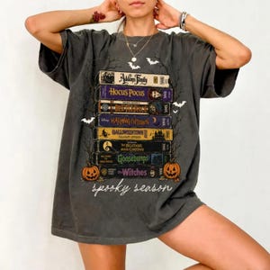 May include: A charcoal gray t-shirt featuring a stack of VHS tapes with titles including The Addams Family, Hocus Pocus, Beetlejuice, Halloweentown, The Nightmare Before Christmas, Goosebumps, and The Witches, with two orange pumpkins and text.