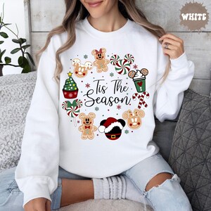 May include: A white crewneck sweatshirt featuring a circular graphic of holiday-themed icons including gingerbread characters, peppermint candies, a Christmas tree cupcake, and a Santa hat. The text 'Tis The Season' is printed in black script.