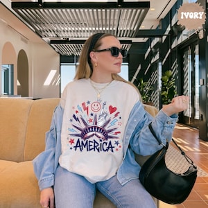 Peut inclure: Un sweat-shirt &agrave; col rond ivoire avec un motif repr&eacute;sentant la Statue de la Libert&eacute;, un visage souriant, un c&oelig;ur et des &eacute;clairs en rouge, blanc et bleu. Le mot AMERICA est imprim&eacute; en lettres bleues grasses au bas du motif.