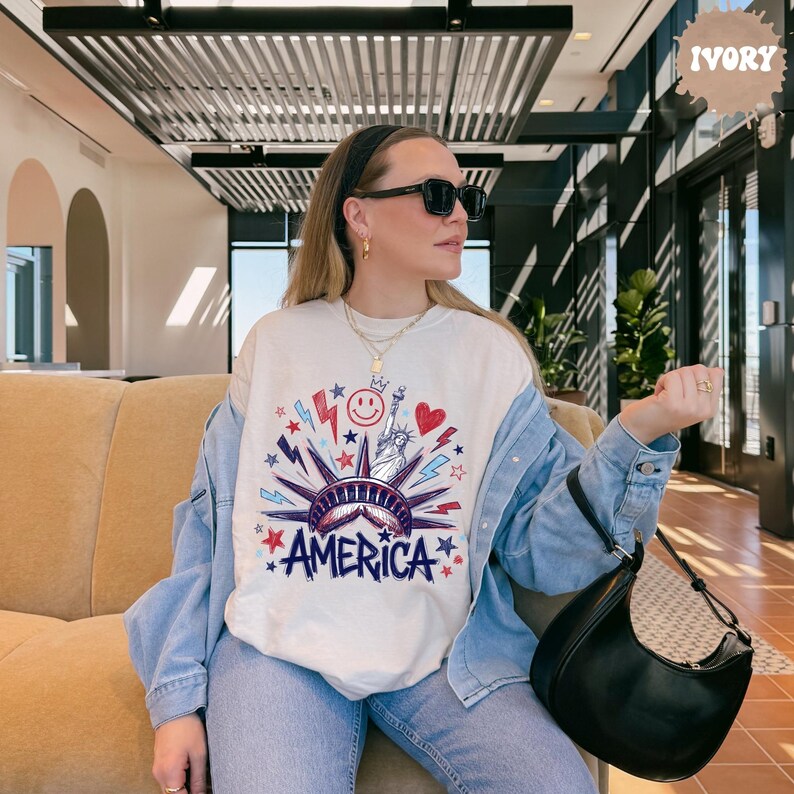 Peut inclure: Un sweat-shirt &agrave; col rond ivoire avec un motif repr&eacute;sentant la Statue de la Libert&eacute;, un visage souriant, un c&oelig;ur et des &eacute;clairs en rouge, blanc et bleu. Le mot AMERICA est imprim&eacute; en lettres bleues grasses au bas du motif.