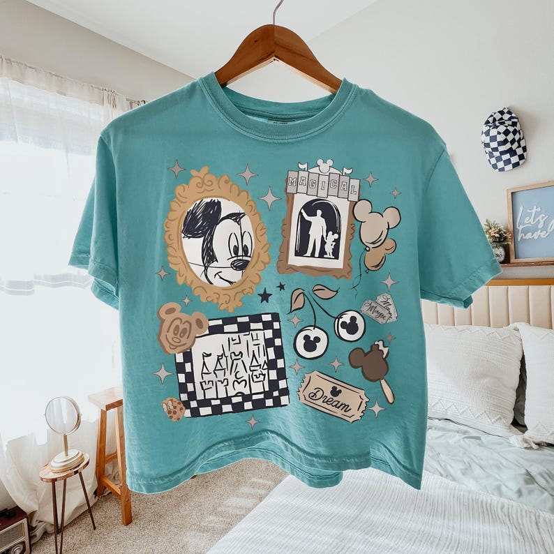 May include: A teal short-sleeve t-shirt featuring a graphic print with a gold-framed Mickey Mouse sketch, a castle in a black and white checkered frame, a ticket labeled Dream, cherry icons with mouse ears, and the word Magical in a banner.