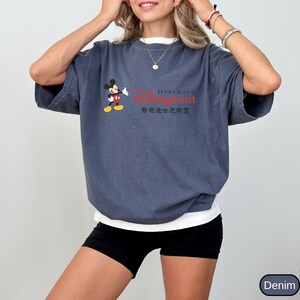 Op de afbeelding: Een denim blauw t-shirt met een wit ondershirt met een Mickey Mouse-afbeelding en de tekst "Hong Kong Disneyland" in het rood en Chinese karakters. Het shirt heeft korte mouwen.