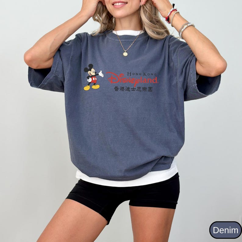 Op de afbeelding: Een denim blauw t-shirt met een wit ondershirt met een Mickey Mouse-afbeelding en de tekst "Hong Kong Disneyland" in het rood en Chinese karakters. Het shirt heeft korte mouwen.