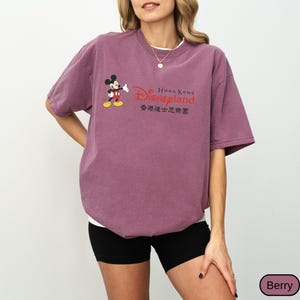 Op de afbeelding: Bessenkleurig T-shirt met Mickey Mouse en het Disneyland Hong Kong logo. Het ontwerp bevat het Disneyland logo in rood en de tekst "Hong Kong Disneyland" in het Engels en Chinees. Het shirt heeft korte mouwen en een ronde hals.