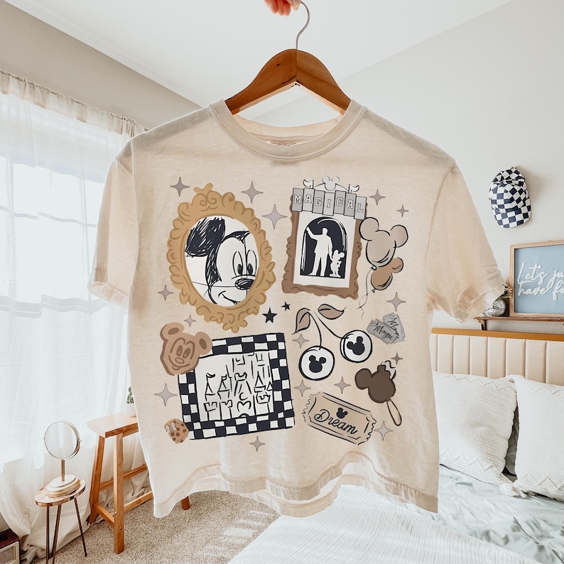 May include: A cream-colored short-sleeve t-shirt featuring a graphic print with a gold-toned mirror frame containing a Mickey Mouse sketch, a sign reading MAGICAL, a checkered square with a castle, cherries, balloons, and a ticket labeled Dream.