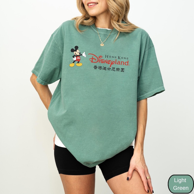 Op de afbeelding: Lichtgroen T-shirt met Mickey Mouse en de tekst "Hong Kong Disneyland" in rood en zwart. Het shirt heeft een ronde hals en korte mouwen. Het shirt is lichtgroen.