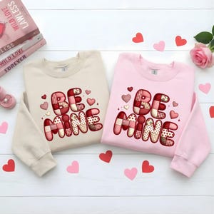 May include: Two sweatshirts, one beige and one pink, are displayed. Each features the phrase "BE MINE" in a patchwork design with hearts. Scattered paper hearts and a rose add to the Valentine's Day theme.