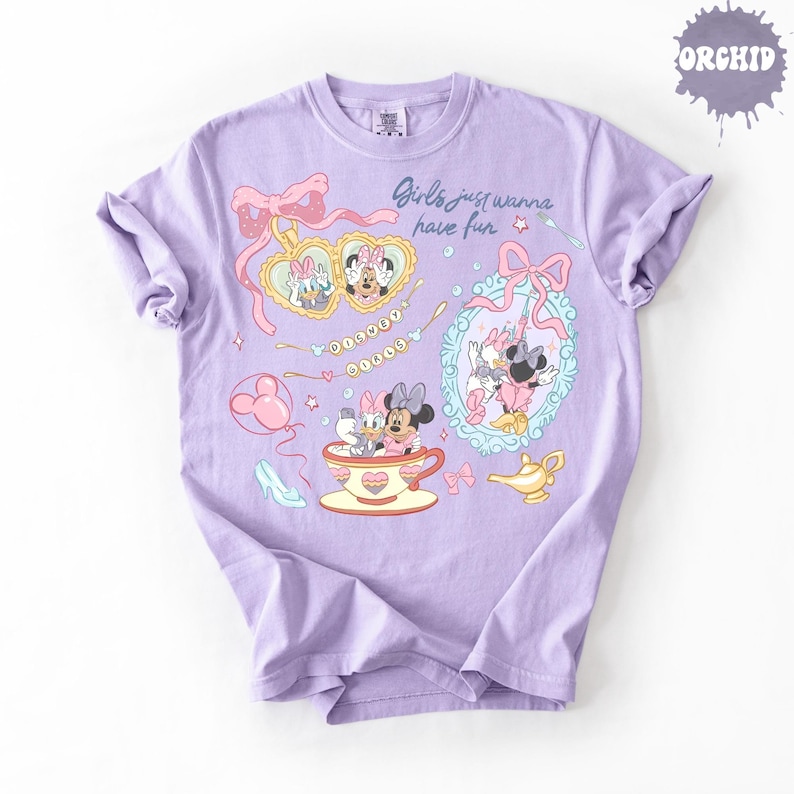 Może przedstawiać: Jasnofioletowy t-shirt z nadrukiem graficznym przedstawiającym Myszkę Minnie i Kaczorkę Daisy. Wz&oacute;r zawiera tekst "Girls just wanna have fun" i "DISNEY GIRLS" oraz ilustracje medalionu w kształcie serca, filiżanki i lustra.