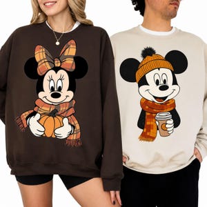 May include: A dark brown sweatshirt featuring a graphic of Minnie Mouse wearing a plaid bow and scarf while holding a pumpkin, and a beige sweatshirt featuring a graphic of Mickey Mouse wearing an orange beanie and scarf while holding a coffee cup.