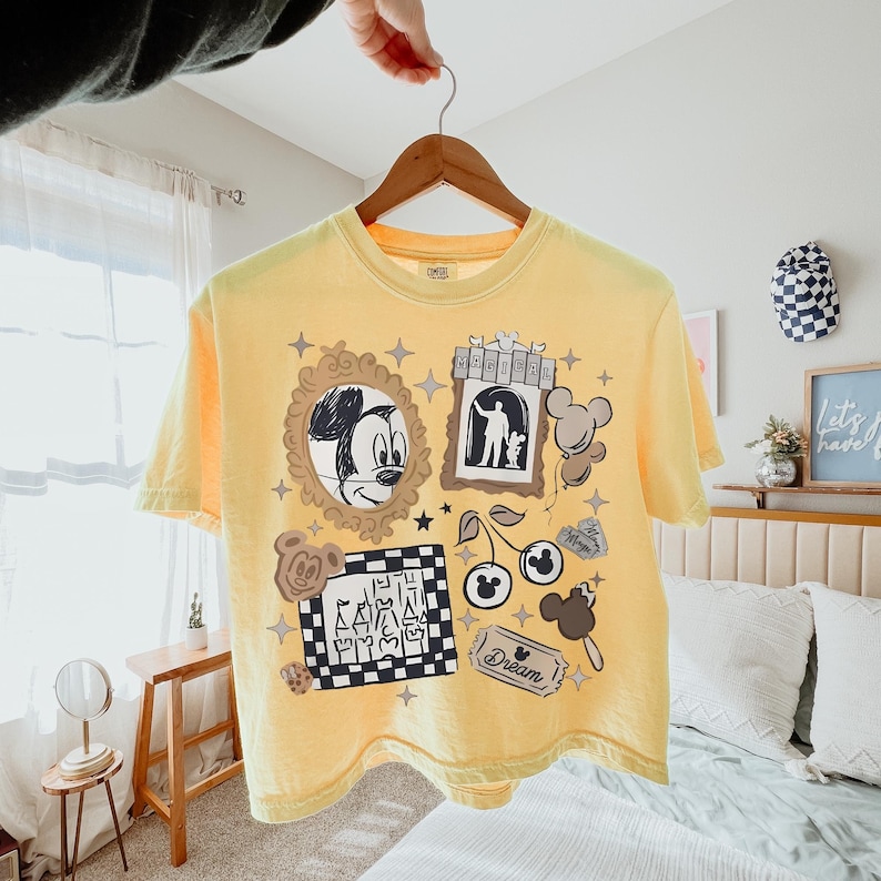 May include: A pale yellow short-sleeve t-shirt featuring a graphic print with a sketch of Mickey Mouse in a gold frame, a castle silhouette on a checkered square, a cherry icon, a ticket labeled Dream, and the word MAGICAL in a banner.