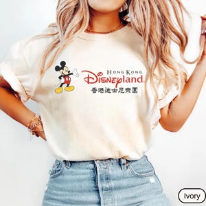 Op de afbeelding: Ivoor kleurig T-shirt met Mickey Mouse en de tekst "Hong Kong Disneyland" in het Engels en Chinees. Mickey Mouse is afgebeeld in zijn klassieke pose. Het shirt heeft een lichtcr&egrave;mekleur.