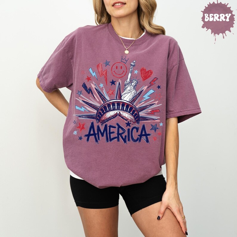 Peut inclure: Un t-shirt &agrave; manches courtes couleur baie avec une impression graphique de la Statue de la Libert&eacute;, le mot AMERICA en bleu, ainsi que des &eacute;clairs, des &eacute;toiles, un c&oelig;ur et un visage souriant rouges et bleus centr&eacute;s sur la poitrine.