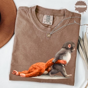 May include: Espresso-colored Comfort Colors t-shirt featuring a graphic of two monkeys. One monkey is gray and the other is orange. A silver necklace with a pendant is draped across the shirt.