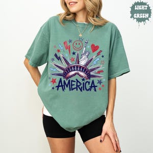 Peut inclure: Un t-shirt vert clair avec une impression graphique de la Statue de la Libert&eacute;, le mot AMERICA, un smiley, un c&oelig;ur et des &eacute;clairs en rouge, blanc et bleu. Le motif comprend diverses formes d'&eacute;toiles dispers&eacute;es autour de la figure centrale.