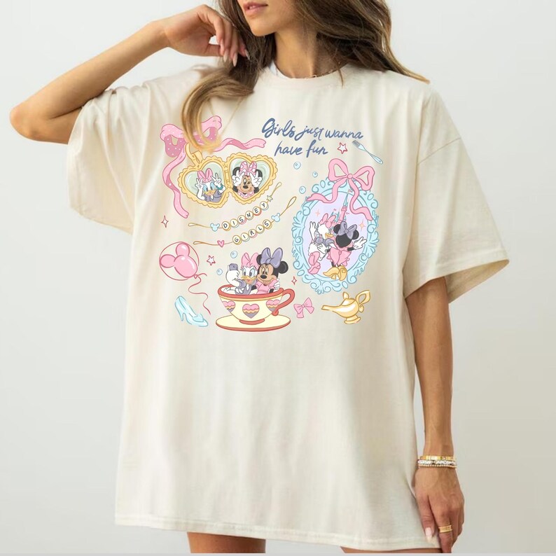 Może przedstawiać: Oversizowy t-shirt w kolorze kremowym z nadrukiem graficznym i napisem "Girls just wanna have fun". Wz&oacute;r zawiera ilustracje Myszki Minnie i Kaczki Daisy, lustra w kształcie serc, filiżankę, szklany pantofelek oraz złotą lampę.