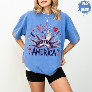 Peut inclure: Un t-shirt bleu &agrave; manches courtes avec une impression graphique de la Statue de la Libert&eacute;, le mot AMERICA, un visage souriant, un c&oelig;ur, ainsi que diverses &eacute;toiles et &eacute;clairs en rouge, blanc et bleu. Le t-shirt a un col rond et une coupe d&eacute;contract&eacute;e.