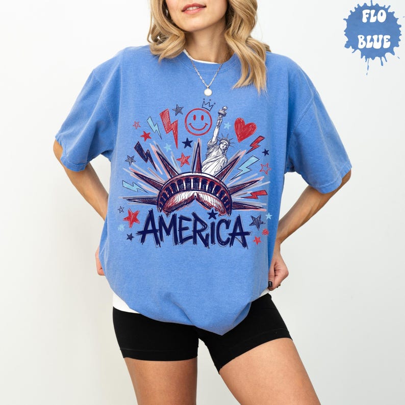 Peut inclure: Un t-shirt bleu &agrave; manches courtes avec une impression graphique de la Statue de la Libert&eacute;, le mot AMERICA, un visage souriant, un c&oelig;ur, ainsi que diverses &eacute;toiles et &eacute;clairs en rouge, blanc et bleu. Le t-shirt a un col rond et une coupe d&eacute;contract&eacute;e.