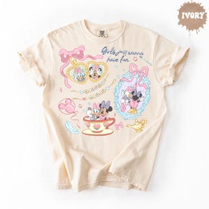 Może przedstawiać: Koszulka w kolorze kości słoniowej z nadrukiem graficznym i napisem "Girls just wanna have fun". Wz&oacute;r zawiera ilustracje Daisy Duck i Minnie Mouse, r&oacute;żowe kokardy, lustro w kształcie serca, filiżankę, magiczną lampę oraz pantofelek.