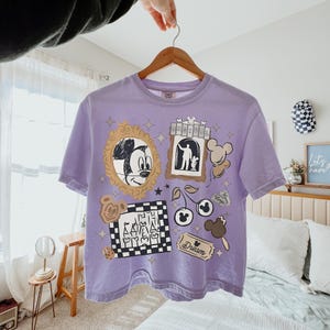 May include: A light purple short-sleeve t-shirt featuring a graphic print with a gold-framed Mickey Mouse sketch, a castle in a checkered frame, a ticket labeled Dream, a cherry icon with Mickey ears, and a sign with the text MAGICAL.