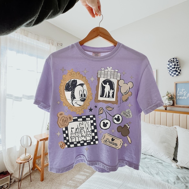 May include: A light purple short-sleeve t-shirt featuring a graphic print with a gold-framed Mickey Mouse sketch, a castle in a checkered frame, a ticket labeled Dream, a cherry icon with Mickey ears, and a sign with the text MAGICAL.