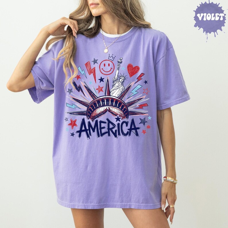 Peut inclure: Un t-shirt &agrave; manches courtes violet clair avec une impression graphique de la Statue de la Libert&eacute;, un smiley, un c&oelig;ur et des &eacute;clairs. Le mot AMERICA est imprim&eacute; en grandes lettres bleu fonc&eacute; sous l'illustration de la Statue de la Libert&eacute;.