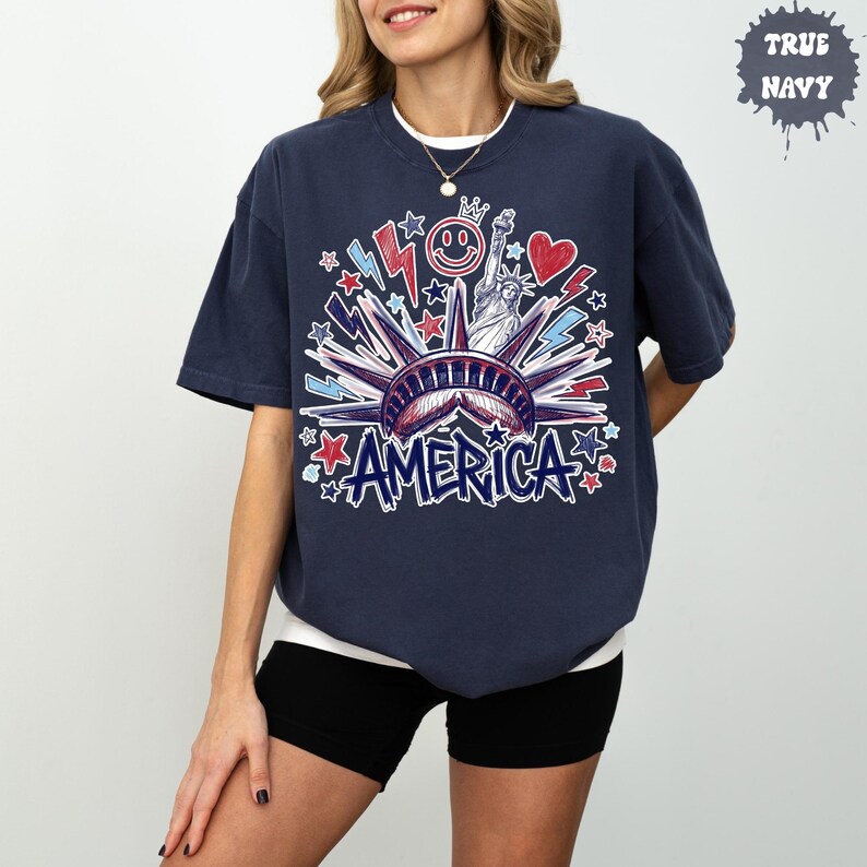 Peut inclure: Un t-shirt bleu marine &agrave; manches courtes avec une impression graphique de la Statue de la Libert&eacute;, un smiley, un c&oelig;ur et des &eacute;clairs en rouge, blanc et bleu. Le mot AMERICA est imprim&eacute; en bleu dans une police stylis&eacute;e en bas du motif.