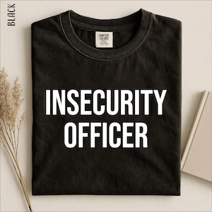 Op de afbeelding: Een zwart t-shirt met ronde hals en de witte tekst INSECURITY OFFICER in een vet, schreefloos lettertype op de borst. Het kledingstuk is voorzien van een Comfort Colors merklabel in de binnenhals en heeft korte mouwen.