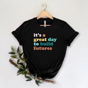 Peut inclure: T-shirt noir avec l'inscription "it's a great day to build futures" en rose, jaune, bleu et orange. Le t-shirt est suspendu &agrave; un cintre en bois sur fond blanc. Des feuilles vertes et une branche brune se trouvent dans le coin inf&eacute;rieur gauche.