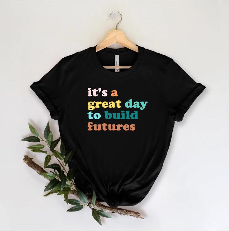 Peut inclure: T-shirt noir avec l'inscription "it's a great day to build futures" en rose, jaune, bleu et orange. Le t-shirt est suspendu &agrave; un cintre en bois sur fond blanc. Des feuilles vertes et une branche brune se trouvent dans le coin inf&eacute;rieur gauche.