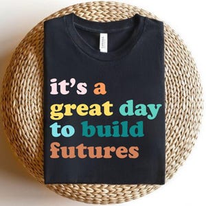 Peut inclure: T-shirt noir avec l'inscription color&eacute;e "it's a great day to build futures" dans un style r&eacute;tro. Le t-shirt est pr&eacute;sent&eacute; sur un support rond en osier.