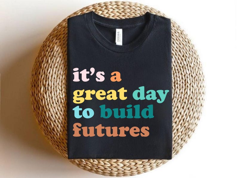 Peut inclure: T-shirt noir avec l'inscription color&eacute;e "it's a great day to build futures" dans un style r&eacute;tro. Le t-shirt est pr&eacute;sent&eacute; sur un support rond en osier.