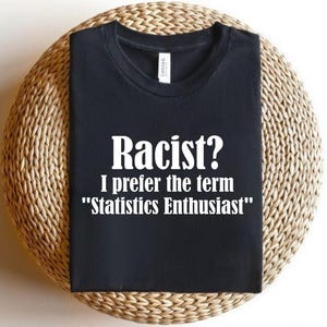 May include: Black t-shirt with white text: "Racist? I prefer the term "Statistics Enthusiast"". The shirt is placed on a circular woven mat.