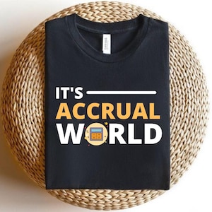 May include: Black t-shirt featuring the phrase "IT'S ACCRUAL WORLD" in white and gold. A calculator graphic is incorporated into the design. The t-shirt is presented on a circular woven mat.