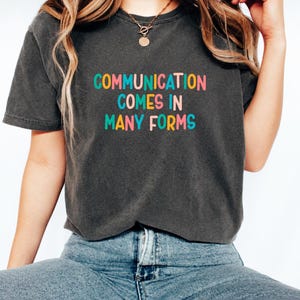 Könnte beinhalten: Ein anthrazitfarbenes Kurzarm-T-Shirt mit dem Text COMMUNICATION COMES IN MANY FORMS in einer mehrfarbigen serifenlosen Schriftart. Das T-Shirt hat einen Rundhalsausschnitt und eine verwaschene Optik, kombiniert mit einer blauen Jeanshose.