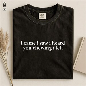 May include: A black crew neck t-shirt featuring the white text "i came i saw i heard you chewing i left" printed on the centre chest. The shirt has a Comfort Colors brand label on the inside of the collar and short sleeves.