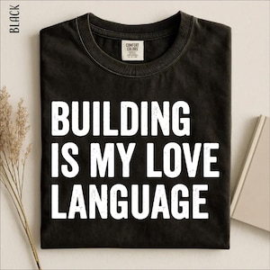 May include: A black short-sleeved t-shirt with a crew neckline featuring the white distressed text "BUILDING IS MY LOVE LANGUAGE" printed on the front. The shirt has a Comfort Colors brand label on the inside of the collar and is labeled as black.