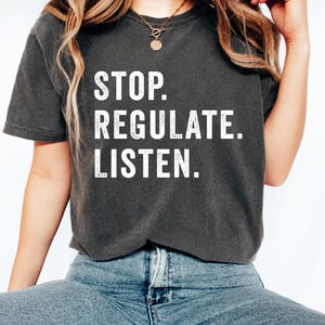 May include: A charcoal grey short-sleeve t-shirt featuring the white distressed-style text STOP. REGULATE. LISTEN. printed in a bold, sans-serif font across the chest. The garment has a crew neckline and a vintage-inspired washed fabric finish.