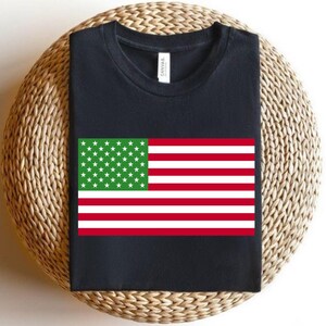 Könnte beinhalten: Ein schwarzes T-Shirt mit Rundhalsausschnitt und einem rechteckigen Flaggenmotiv, das links ein grünes Feld mit weißen Sternen und rechts horizontale rote und weiße Streifen zeigt. Am inneren Halsausschnitt befindet sich ein weißes Markenetikett.