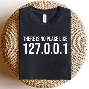 May include: Black t-shirt with the white text "THERE IS NO PLACE LIKE 127.0.0.1". The shirt is placed on a circular woven mat.
